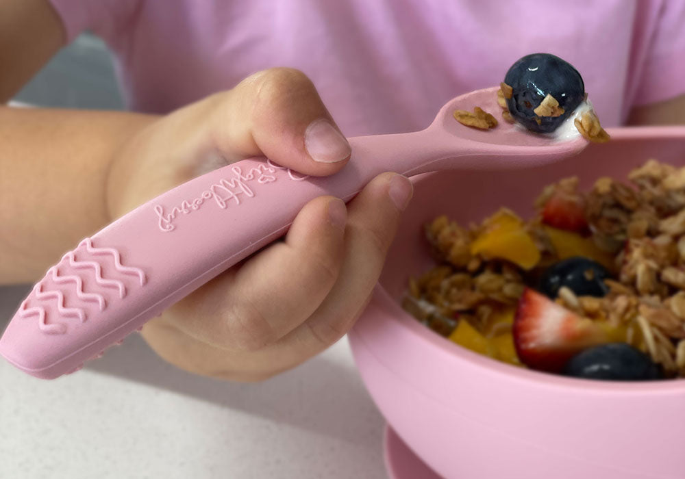 child holding silicone spoon and scooping from suction bowl with suction base and spoon in pink colour